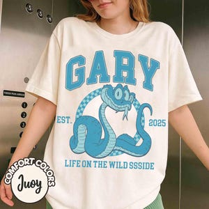 May include: Cream-colored t-shirt with a blue graphic featuring the name "GARY" in a collegiate font above a cartoon snake. The snake is inside a checkered circle, with the text "EST. 2025" and "LIFE ON THE WILD SSSIDE" below.
