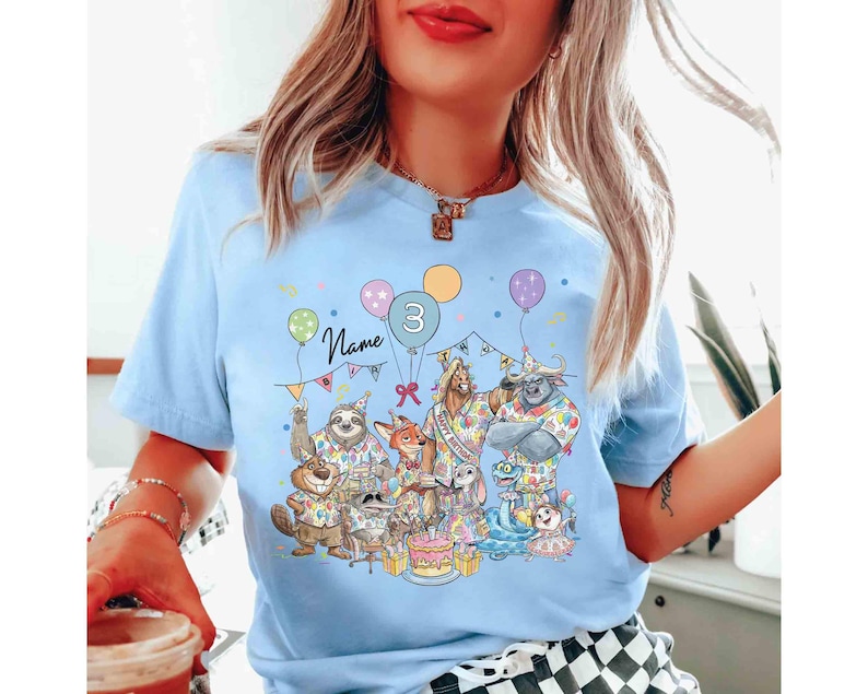 May include: Light blue t-shirt featuring a colorful birthday party design. The graphic includes cartoon animals, balloons, a cake, and the number 3. The word "Name" is written above the balloons. The shirt is a short-sleeved crew neck.