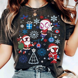 May include: Dark gray t-shirt featuring a Christmas-themed design with cartoon characters dressed as Spider-Man and Spider-Gwen. The design includes snowflakes, ornaments, and a Christmas tree. The characters are wearing Santa hats and red and blue suits.