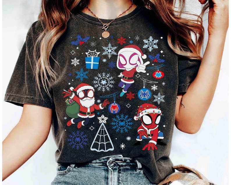 May include: Dark gray t-shirt featuring a Christmas-themed design with cartoon characters dressed as Spider-Man and Spider-Gwen. The design includes snowflakes, ornaments, and a Christmas tree. The characters are wearing Santa hats and red and blue suits.