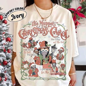 Puede incluir: Camiseta de color marfil con un gráfico de "The Muppet Christmas Carol". El diseño incluye el título, los personajes y frases como "I am here to tell the story" y "I am here for the food". La camiseta es de la marca Comfort Colors.