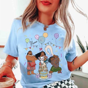 Puede incluir: Camiseta azul claro con un gr&aacute;fico colorido de personajes de dibujos animados, una tarta de cumplea&ntilde;os y globos. Tambi&eacute;n se muestra el texto "Name" y el n&uacute;mero "5". La camiseta es de cuello redondo y manga corta.