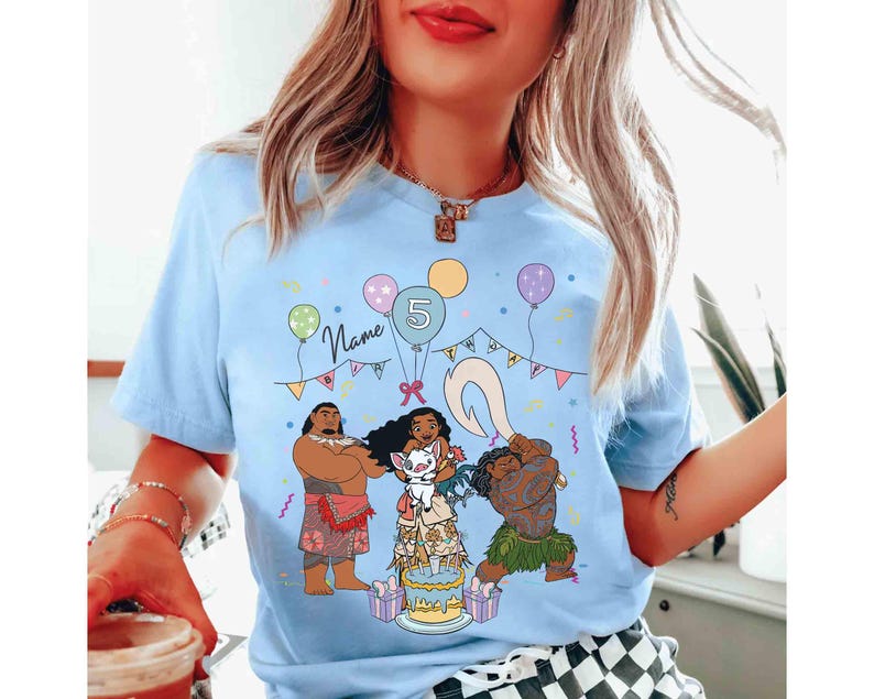 Puede incluir: Camiseta azul claro con un gr&aacute;fico colorido de personajes de dibujos animados, una tarta de cumplea&ntilde;os y globos. Tambi&eacute;n se muestra el texto "Name" y el n&uacute;mero "5". La camiseta es de cuello redondo y manga corta.