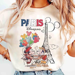 May include: Cream-coloured t-shirt with a graphic of Mickey and Minnie Mouse in Paris. The design includes the Eiffel Tower, balloons, and the text "PARIS Disneyland" in red, white, and blue.