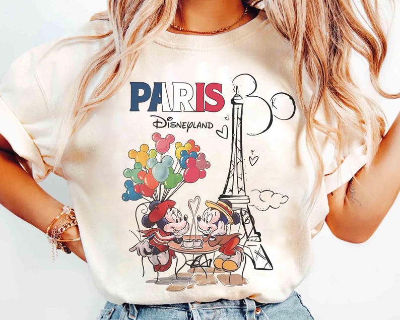 May include: Cream-coloured t-shirt with a graphic of Mickey and Minnie Mouse in Paris. The design includes the Eiffel Tower, balloons, and the text "PARIS Disneyland" in red, white, and blue.