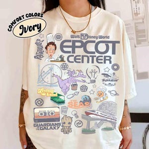 Peut inclure: T-shirt ivoire avec un motif graphique comprenant le texte "EPCOT CENTER" et diverses illustrations sur le th&egrave;me de Disney. Le motif comprend des personnages et des ic&ocirc;nes d'attractions telles que "Guardians of the Galaxy" et "Ratatouille".