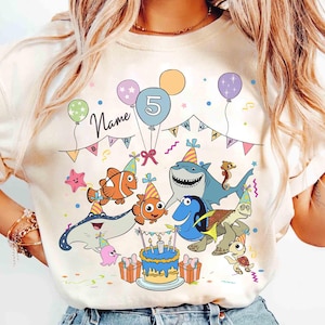 May include: Cream-colored t-shirt featuring a colorful cartoon design of characters from a popular animated film celebrating a fifth birthday. The design includes balloons, a cake, and the number 5. The word "Name" is also visible.