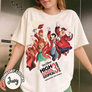 May include: Cream-colored t-shirt featuring a Disney cartoon graphic of Goofy and friends in graduation attire. The text reads "High School Goofical Senior Year" with "Graduating This Spring!" below. The shirt has a Comfort Colors label.