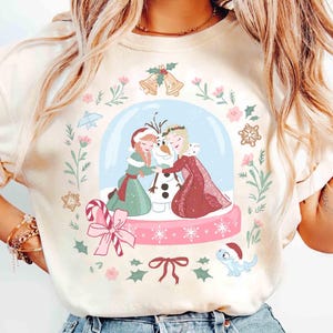 Puede incluir: Sudadera de color crema con un diseño de bola de nieve con dos personajes de dibujos animados, un muñeco de nieve y detalles florales. El diseño incluye elementos rosas, verdes y azules, con un bastón de caramelo y un lazo. El tema general es invierno y festivo.