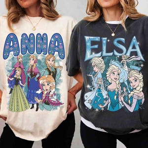 Könnte beinhalten: Zwei T-Shirts mit den Charakteren Anna und Elsa aus dem Film Frozen. Das Anna-Shirt ist cremefarben mit einem blauen Blumendesign um den Namen und Bildern von Anna. Das Elsa-Shirt ist dunkelgrau mit dem Namen Elsa in Blau und Bildern von Elsa.