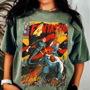 Può includere: T-shirt verde con una copertina di fumetto vintage. Il design mostra un team di supereroi e le parole "The Fantastic", "First Family" e "First Steps". Colori: rosso, giallo e blu.