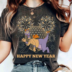 Puede incluir: Camiseta gris oscuro con un personaje de dibujos animados de dragón con una chaqueta amarilla y cuerpo morado, con fuegos artificiales y una esfera geométrica de fondo. El texto "HAPPY NEW YEAR" está impreso en dorado.