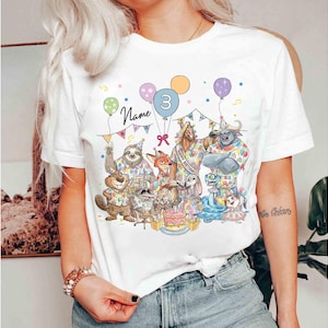 May include: White t-shirt featuring a colorful illustration of cartoon animals celebrating a birthday. The design includes balloons, a cake, and the number 3. The word "Name" is written above the animals.