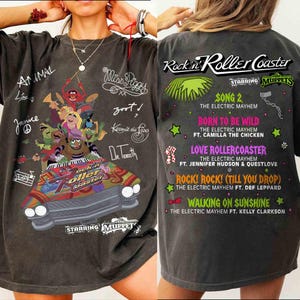 Puede incluir: Camiseta gris oscuro con un gráfico colorido de los Muppets en una montaña rusa, con texto que incluye "Rock 'n' Roller Coaster" y nombres de personajes. La parte trasera de la camiseta enumera títulos de canciones.