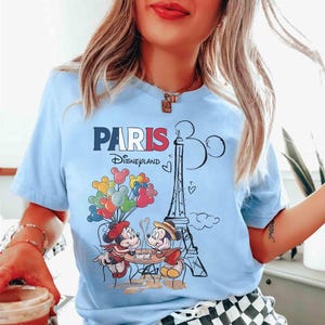 May include: Pale blue t-shirt with a graphic of Mickey and Minnie Mouse in Paris, France. The design includes the Eiffel Tower, balloons, and the text "PARIS Disneyland". The shirt has short sleeves.