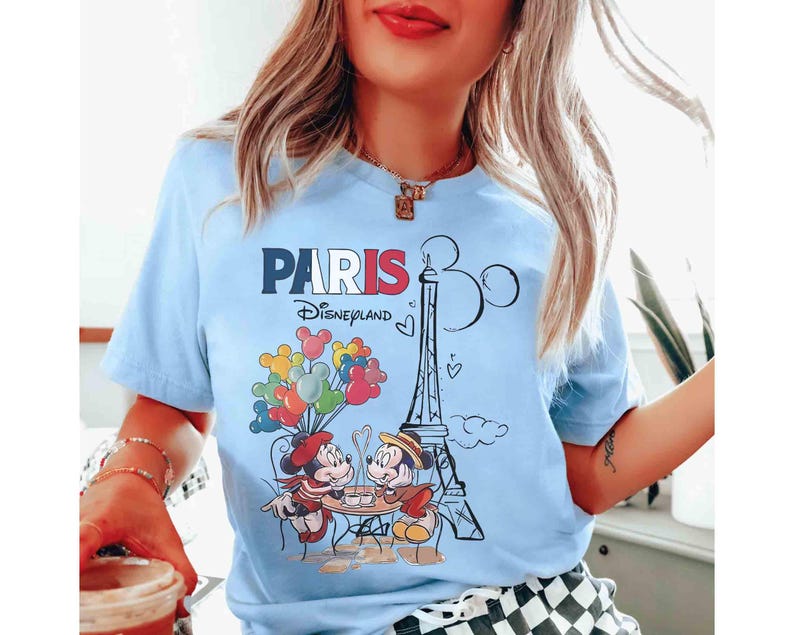 May include: Pale blue t-shirt with a graphic of Mickey and Minnie Mouse in Paris, France. The design includes the Eiffel Tower, balloons, and the text "PARIS Disneyland". The shirt has short sleeves.