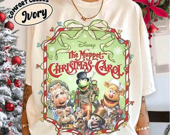 Op de afbeelding: Ivoor kleurig T-shirt met een afbeelding van Disney's "The Muppet Christmas Carol". Het ontwerp bevat Kermit de Kikker, Miss Piggy en andere Muppet-personages in een groen en rood kader. Op het shirt staan de woorden "Comfort Colors" en "Ivory".