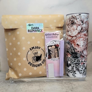 May include: A brown paper bag with white polka dots, a sticker that says "Blind Date with a Book Dark Romance", a sticker with a skeleton couple and the text "Dark Romance", a sticker with the text "Alphabet Challenge", a purple and white gridded bookmark with the text "Reader", a purple pen, and a white tumbler with a floral design and the text "Morally Grey Book Club".