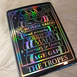 May include: A black phone case with a holographic design featuring a list of popular romance tropes, including "Fake Dating", "One Bed", "Reverse Harem", "Forced Proximity", "Morally Grey", "Mafia", "Enemies to Lovers", "Age Gap", and "The Tropes".