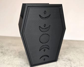 May include: A black, coffin-shaped container with a raised design of crescent moon phases. The container has a matte finish and is set against a neutral background. The design is a decorative element.