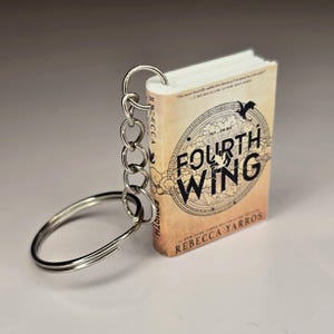 May include: Miniature book keychain titled "Fourth Wing" by Rebecca Yarros. The book has a tan cover with black lettering and a circular design. The keychain is attached with silver metal rings.