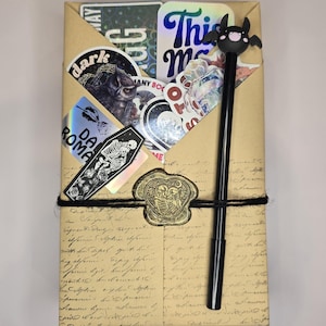 May include: A wrapped package with stickers, a bat-topped pen, and a wax seal. The stickers feature gothic and dark themes, including a coffin and a bat. The pen is black, and the wax seal is heart-shaped with skeleton figures.