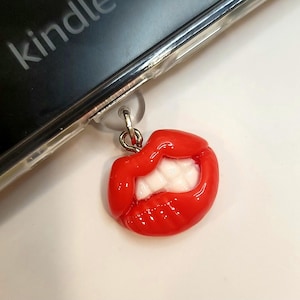 May include: Red plastic charm shaped like a pair of lips with white teeth, attached to a silver chain. The charm is hanging from a clear plastic phone case.