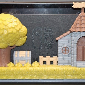 May include: A 3D-printed video game console stand featuring a whimsical landscape with a tree, treasure chest, castle, and fence. The stand is designed to hold a gaming console, with pink and green controllers visible in the background.