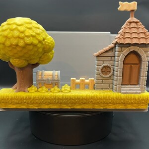 May include: A 3D-printed diorama featuring a yellow tree, a treasure chest, a small fence, and a castle with a flag. The base is yellow, resembling grass. The castle has a brown door and roof.