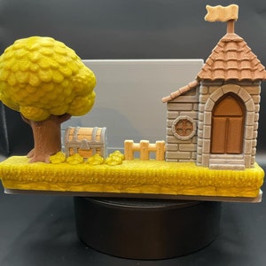 May include: A 3D-printed diorama featuring a castle with a flag, a tree, a treasure chest, and a small fence. The tree has a yellow-green canopy, and the castle has a brown roof and door. The base is yellow, and the overall design is whimsical.