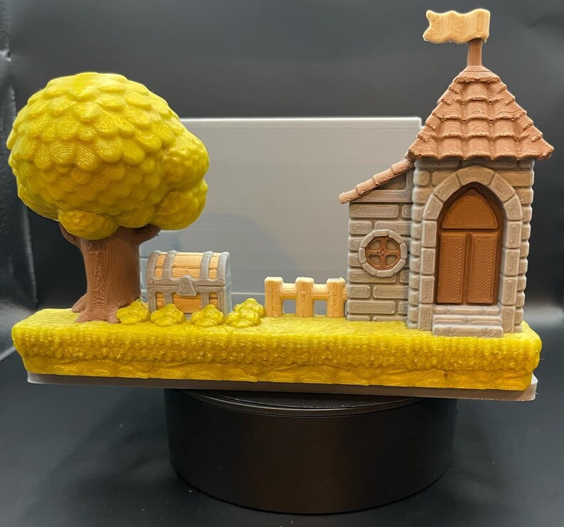 May include: A 3D-printed diorama featuring a castle with a flag, a tree, a treasure chest, and a small fence. The tree has a yellow-green canopy, and the castle has a brown roof and door. The base is yellow, and the overall design is whimsical.