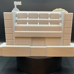 May include: A light beige, 3D-printed model of a building with a flag on top. The model has a rectangular shape with multiple rectangular openings and a layered design. The overall aesthetic is minimalist and architectural.