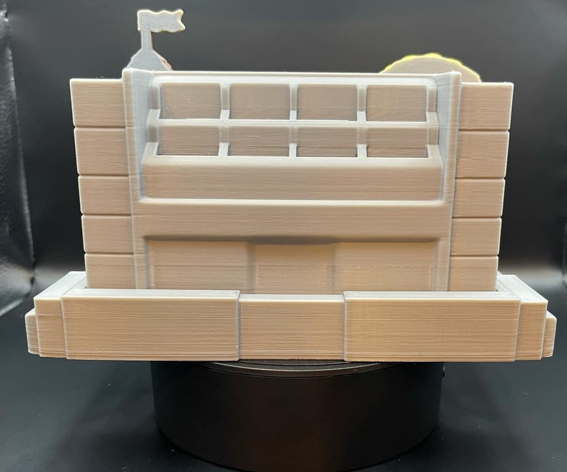 May include: A light beige, 3D-printed model of a building with a flag on top. The model has a rectangular shape with multiple rectangular openings and a layered design. The overall aesthetic is minimalist and architectural.