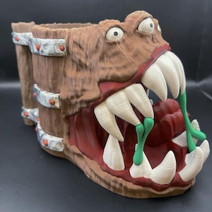 May include: A brown, monster-themed planter with a wide open mouth revealing large white teeth and green drool. The planter has a wooden barrel design with metal bands and red rivets. The eyes are white with black pupils.