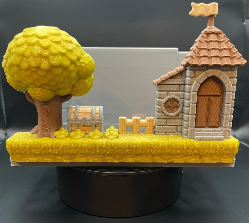 May include: A 3D-printed diorama featuring a yellow tree, a treasure chest, a small fence, and a castle with a flag. The base is yellow, resembling grass. The castle has a brown door and roof.