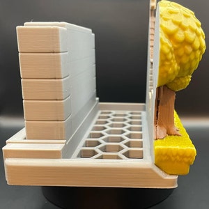 May include: A 3D-printed model of a diorama, featuring a gray building facade, a honeycomb-patterned base, and a tree with yellow foliage. The tree has a brown trunk and sits on a yellow grassy area.