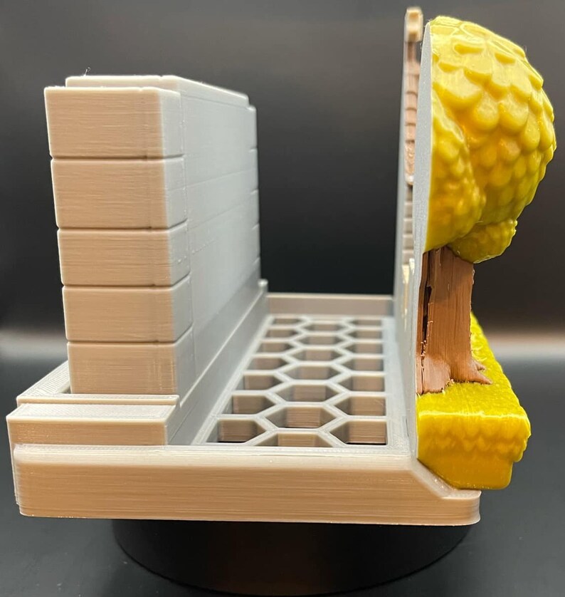 May include: A 3D-printed model of a diorama, featuring a gray building facade, a honeycomb-patterned base, and a tree with yellow foliage. The tree has a brown trunk and sits on a yellow grassy area.
