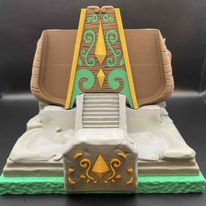 May include: A 3D-printed model of a temple or shrine, featuring a brown roof, a green and gold decorative facade, and a stone base with a staircase. The model has a detailed design with intricate patterns and a green base.