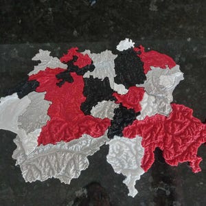 Puede incluir: Un mapa de Suiza impreso en 3D, con detalles topográficos. Los cantones están representados en rojo, negro, blanco y gris. El mapa se encuentra sobre una superficie oscura y texturizada, resaltando los detalles.
