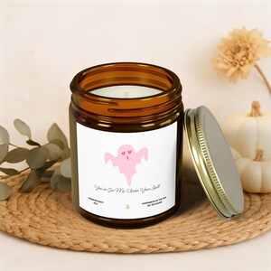 May include: A brown glass jar candle with a metal lid, featuring a pink ghost graphic with heart eyes. The label reads "You've Got Me Under Your Spell." The candle is on a woven mat with a white pumpkin and dried flower in the background.