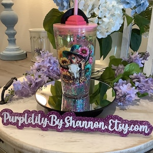 May include: A clear tumbler with a pink lid and straw, featuring a colorful graphic of a woman in a hat with sunflowers and a skull. The tumbler sits on a mirrored surface, with a glittery purple sign that reads "Purplelily By Shannon. Etsy.com".