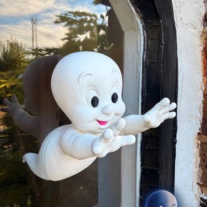 Könnte beinhalten: Eine weiße Casper-Figur mit glatter Oberfläche, großen schwarzen Augen und einem kleinen rosa Mund. Die Figur ist in einer Flugpose mit nach vorne ausgestreckten Armen dargestellt und zeigt den Schriftzug CASPER in blauen und weißen Buchstaben.