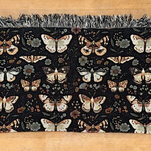 May include: Black woven throw blanket with a repeating pattern of butterflies and floral designs. The blanket has a fringed edge.