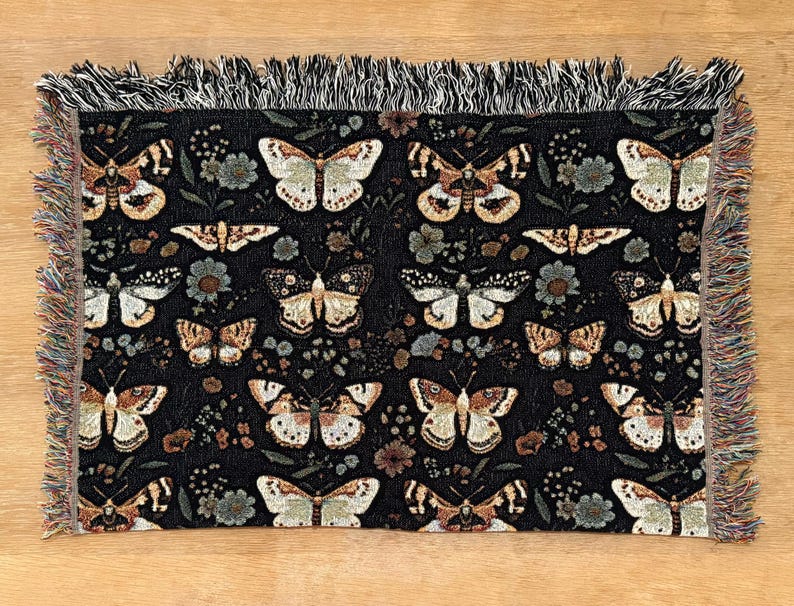 May include: Black woven throw blanket with a repeating pattern of butterflies and floral designs. The blanket has a fringed edge.