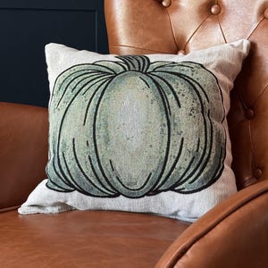 Pode incluir: Uma almofada decorativa quadrada com um grande design de abóbora verde estilizada. A abóbora tem contornos pretos e uma aparência texturizada. A almofada está em uma poltrona de couro marrom.