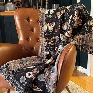 May include: A black and brown woven throw blanket with a butterfly pattern draped over a brown leather armchair. The blanket has a fringe border.