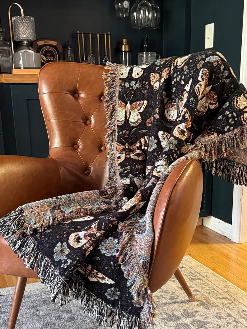 May include: A black and brown woven throw blanket with a butterfly pattern draped over a brown leather armchair. The blanket has a fringe border.