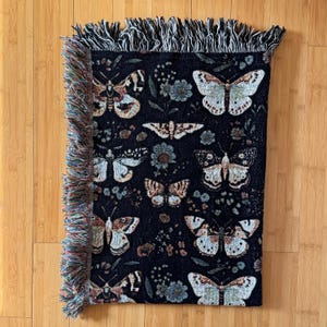 May include: Black woven throw blanket with a fringe border featuring a repeating pattern of butterflies and floral designs.