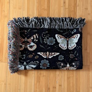 May include: Black woven throw blanket with a fringe border featuring a repeating pattern of butterflies and floral designs.