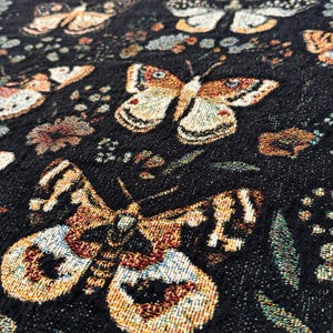May include: Black fabric with a repeating pattern of gold and brown butterflies and floral designs. The fabric has a textured woven look.